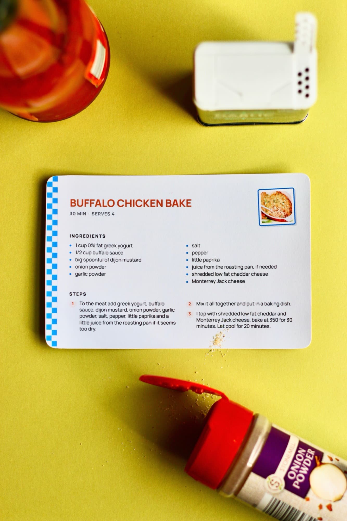 A printed Buffalo Chicken Bake card with a blue checkered edge, on a yellow surface between a jar of buffalo sauce and a tipped-over jar of onion powder.