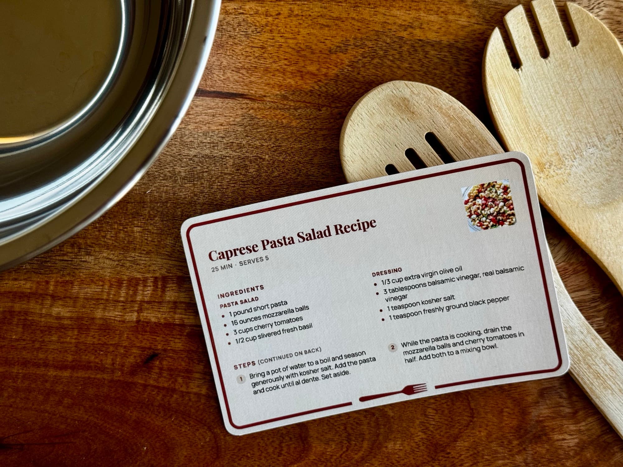 A printed Caprese Pasta Salad card with a burgundy border on a wooden table, between a steel mixing bowl and two wooden serving spoons.
