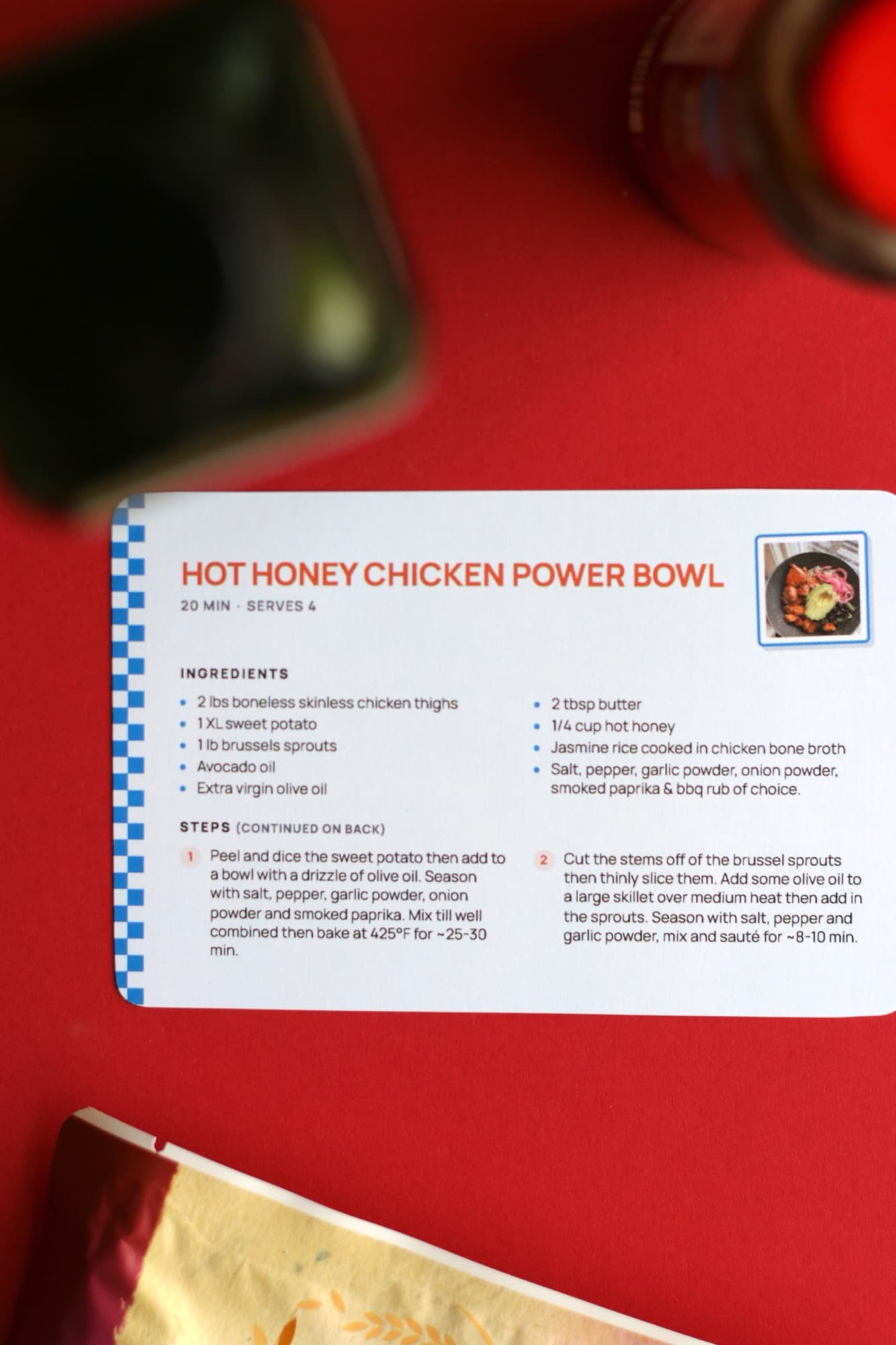 A printed Hot Honey Chicken Power Bowl card with a blue checkered edge, on a red surface beside an avocado.