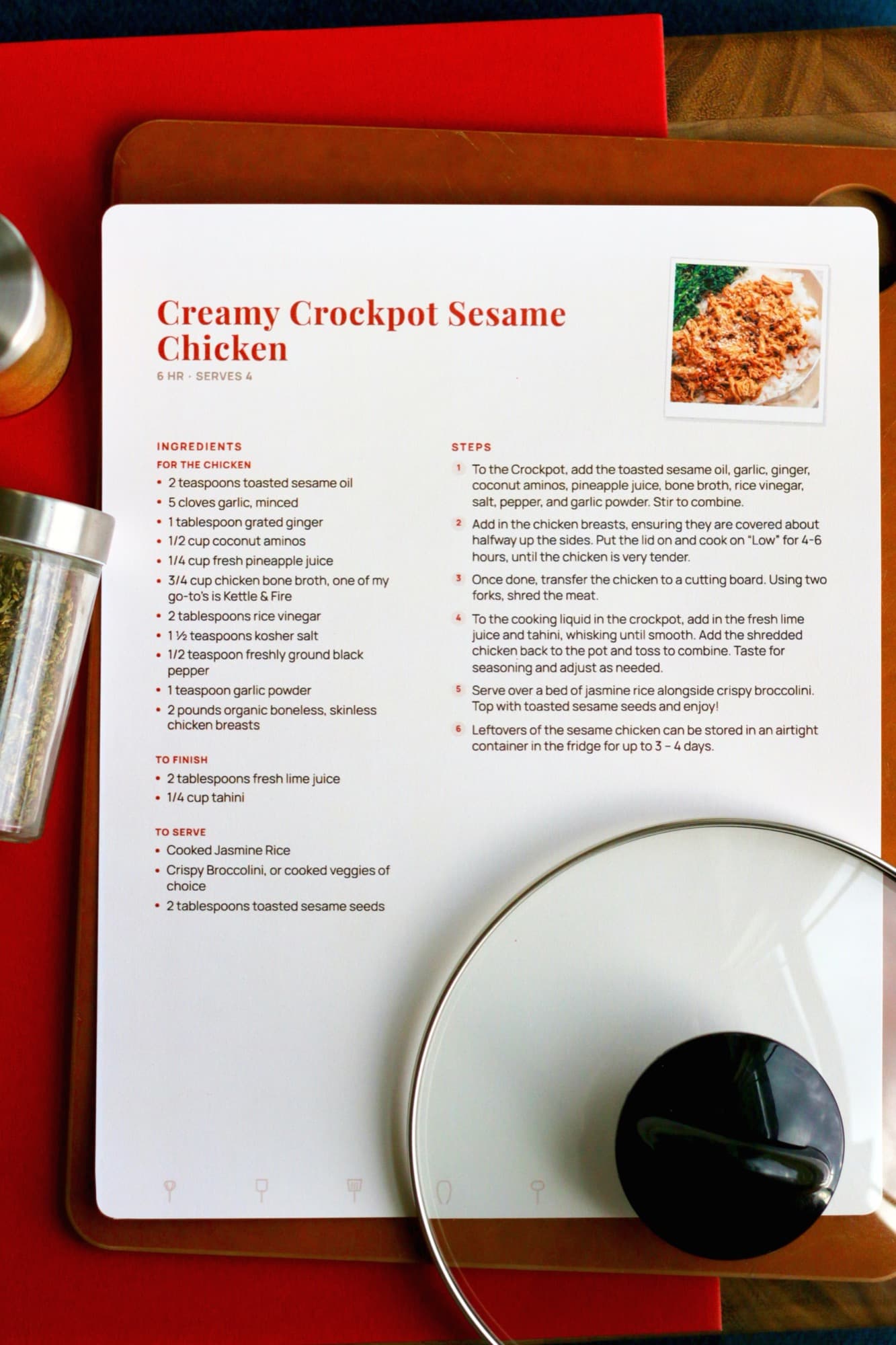 A printed full-page Creamy Crockpot Sesame Chicken recipe on a wooden board, with a glass pot lid resting across one corner.