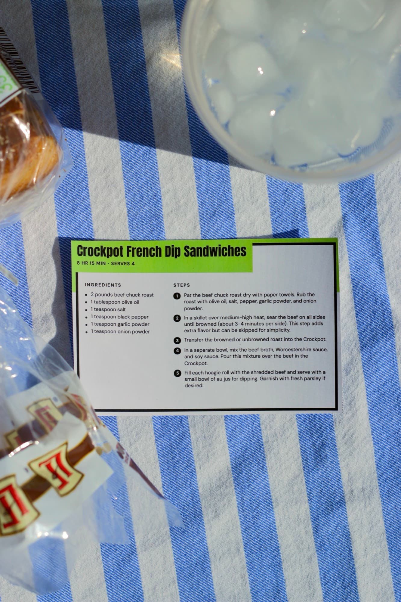 A printed Crockpot French Dip Sandwiches card with a lime-green title band, on a blue-and-white striped towel beside a bag of hoagie rolls and a glass of ice.