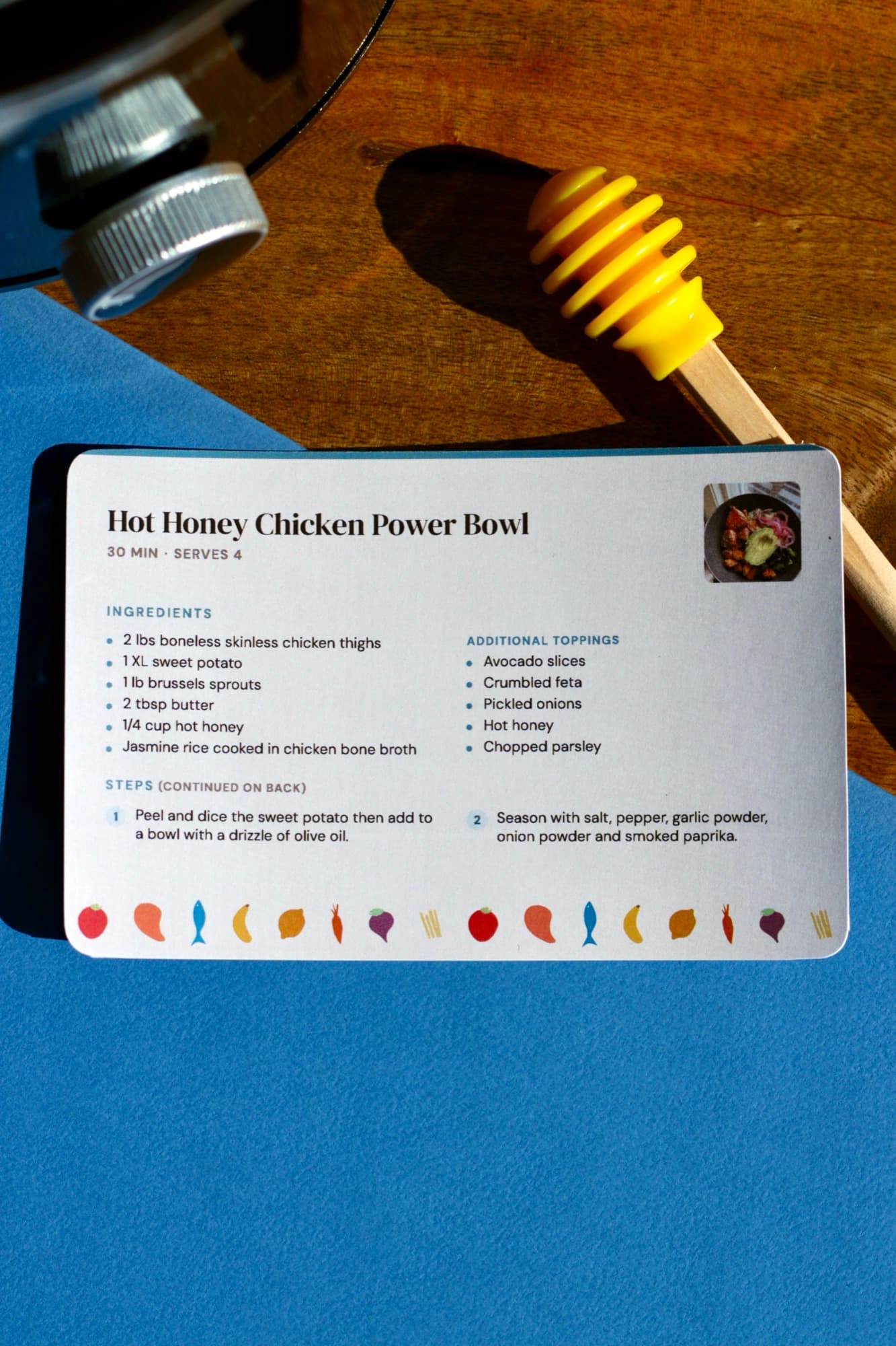 A printed Hot Honey Chicken Power Bowl card with a border of painted vegetables, on a blue surface beside a yellow honey dipper.