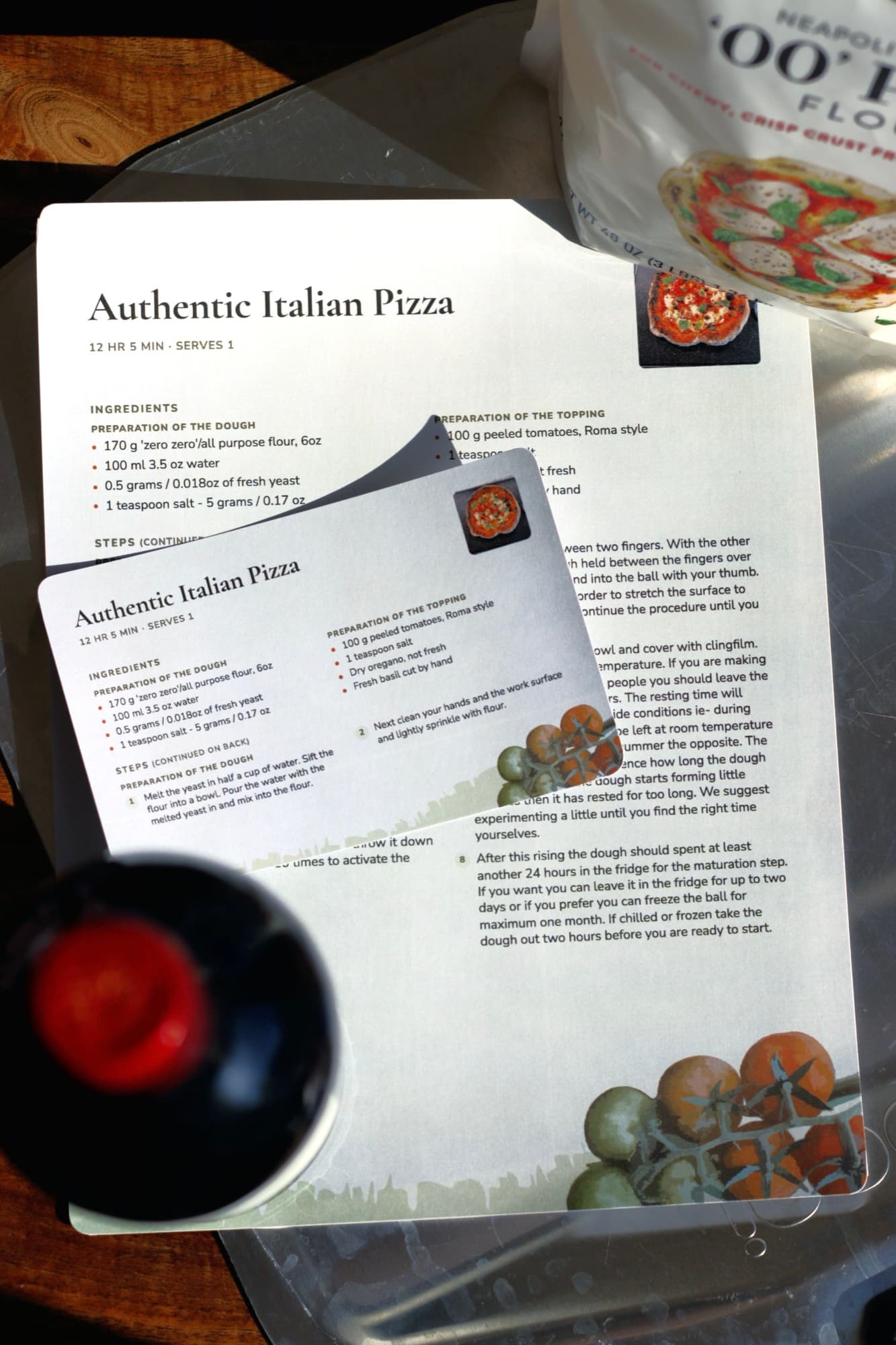 A printed Authentic Italian Pizza card resting on the same recipe printed as a full page, beside a bottle and a bag of 00 flour.