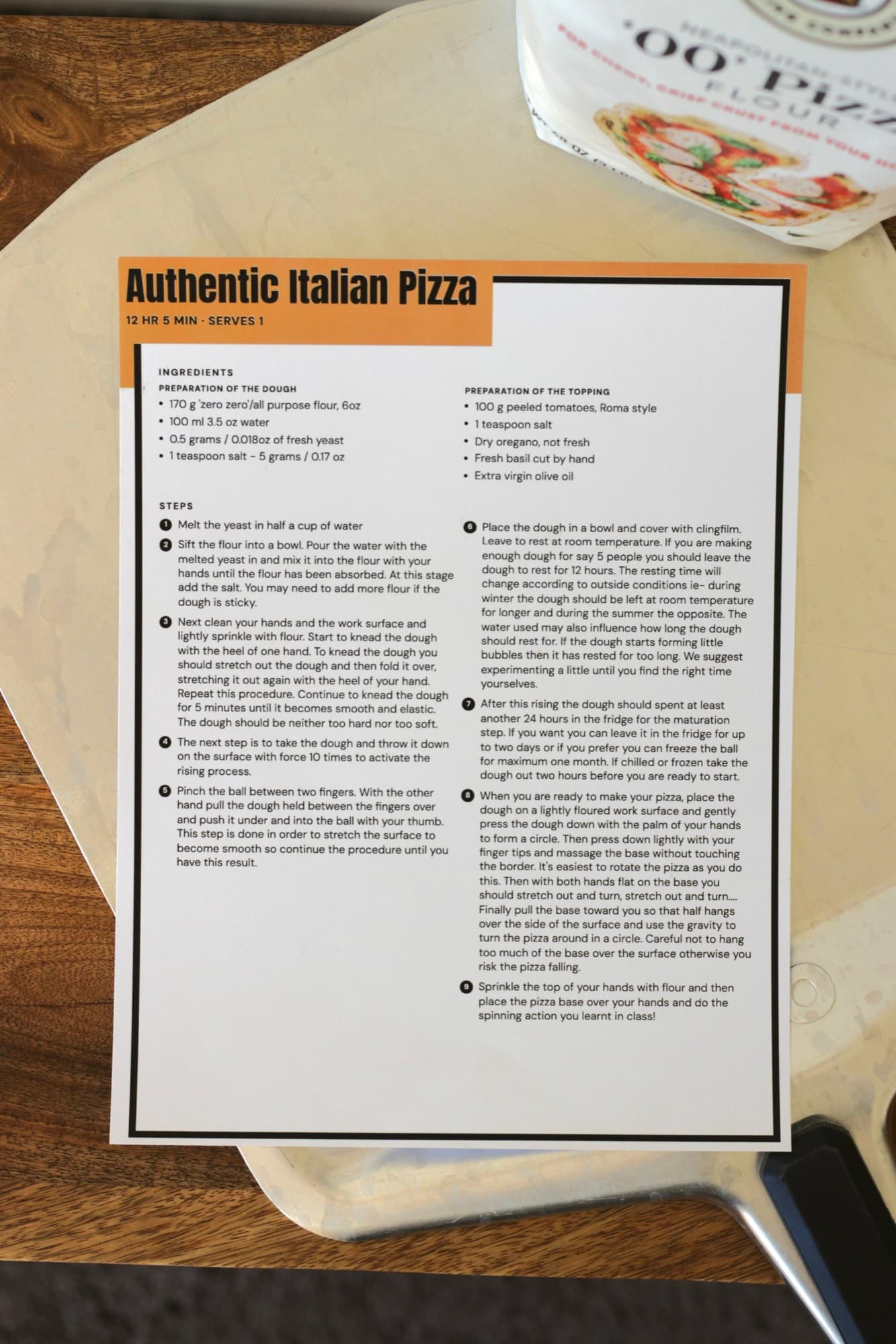 A printed full-page Authentic Italian Pizza recipe with an orange title band, on a pizza peel beside a bag of 00 flour.