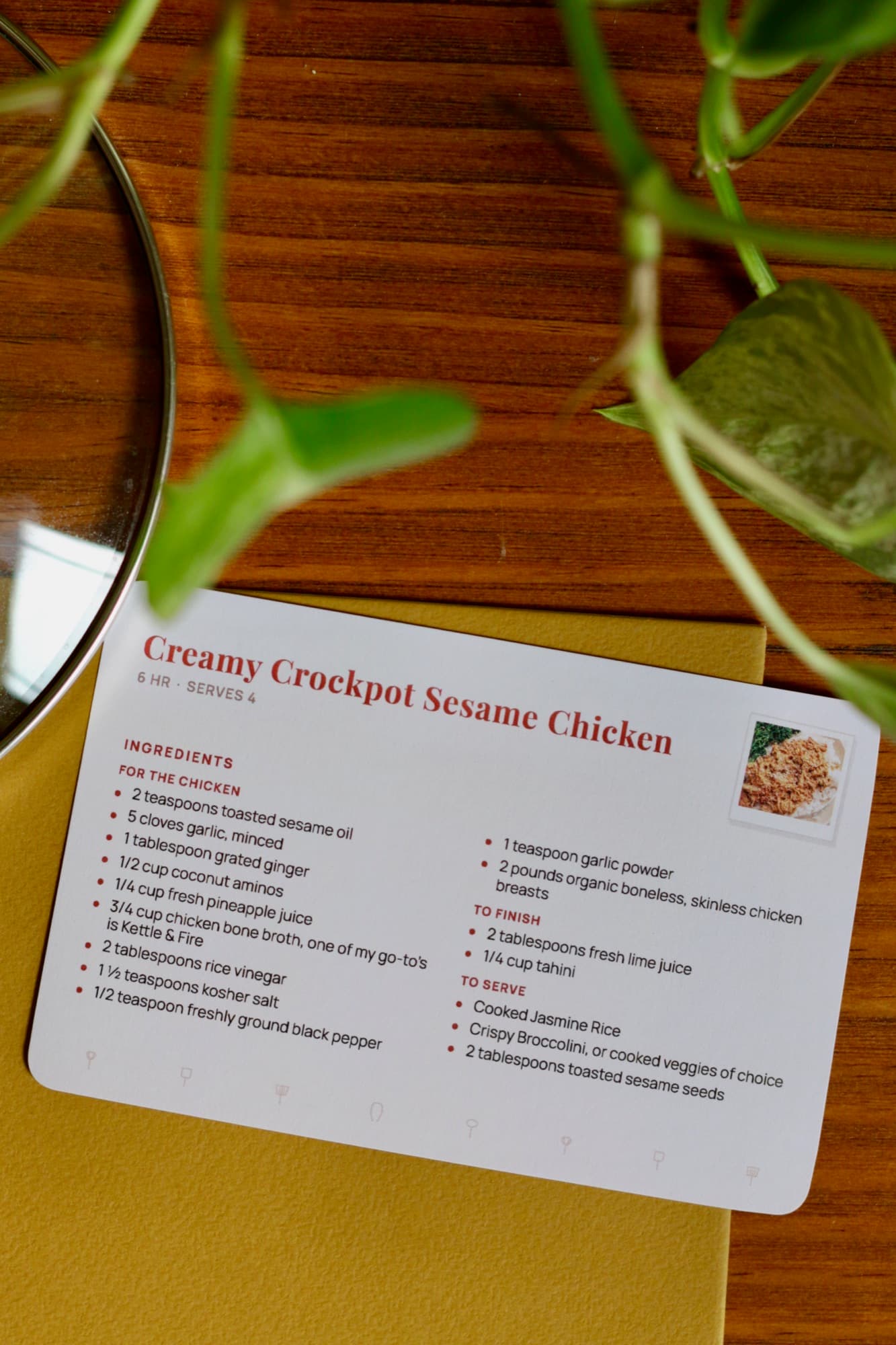 A printed Creamy Crockpot Sesame Chicken card on a mustard notebook, with a trailing houseplant above it.