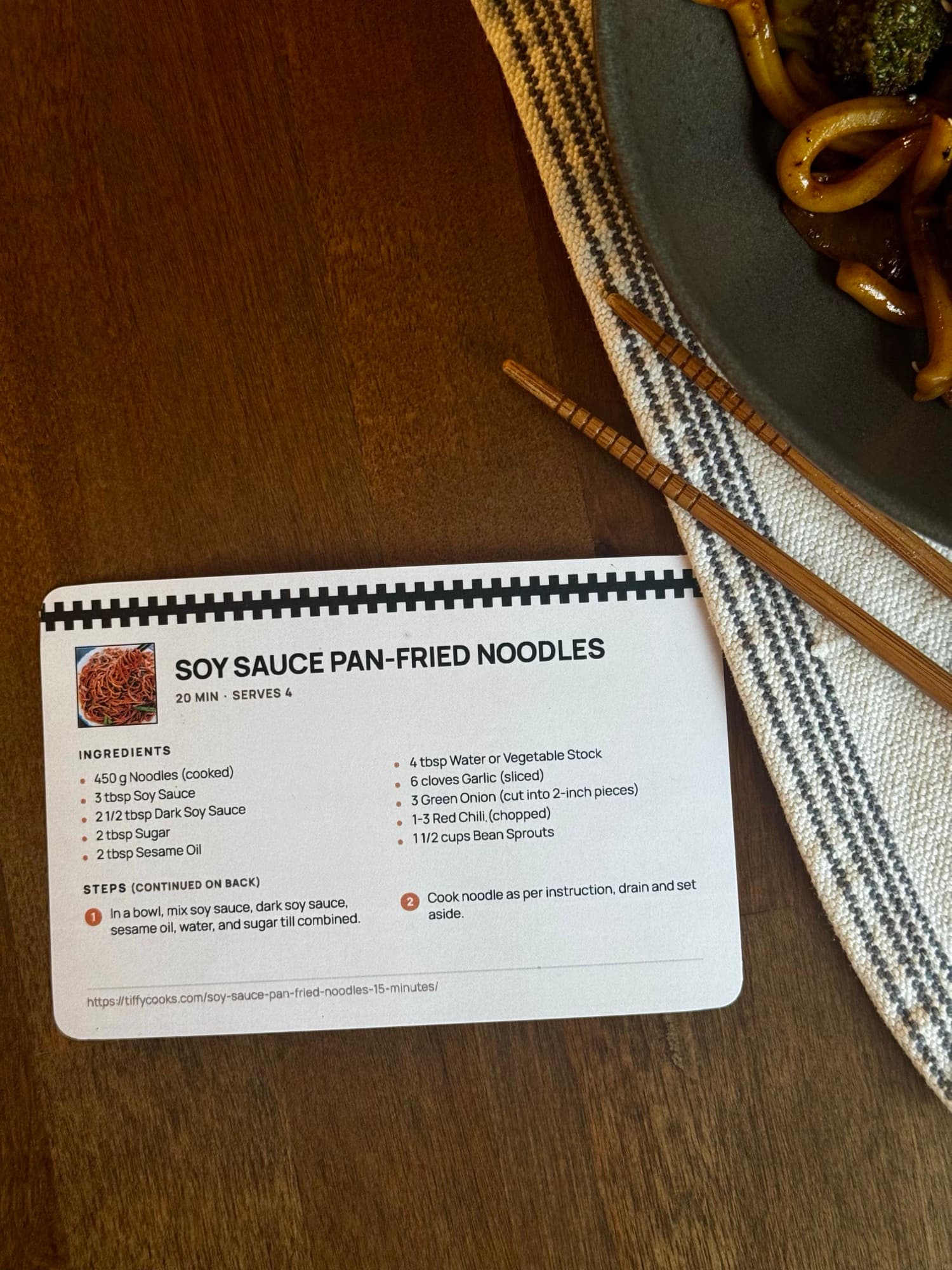 A printed Soy Sauce Pan-Fried Noodles card on a table beside the finished bowl of noodles and chopsticks.