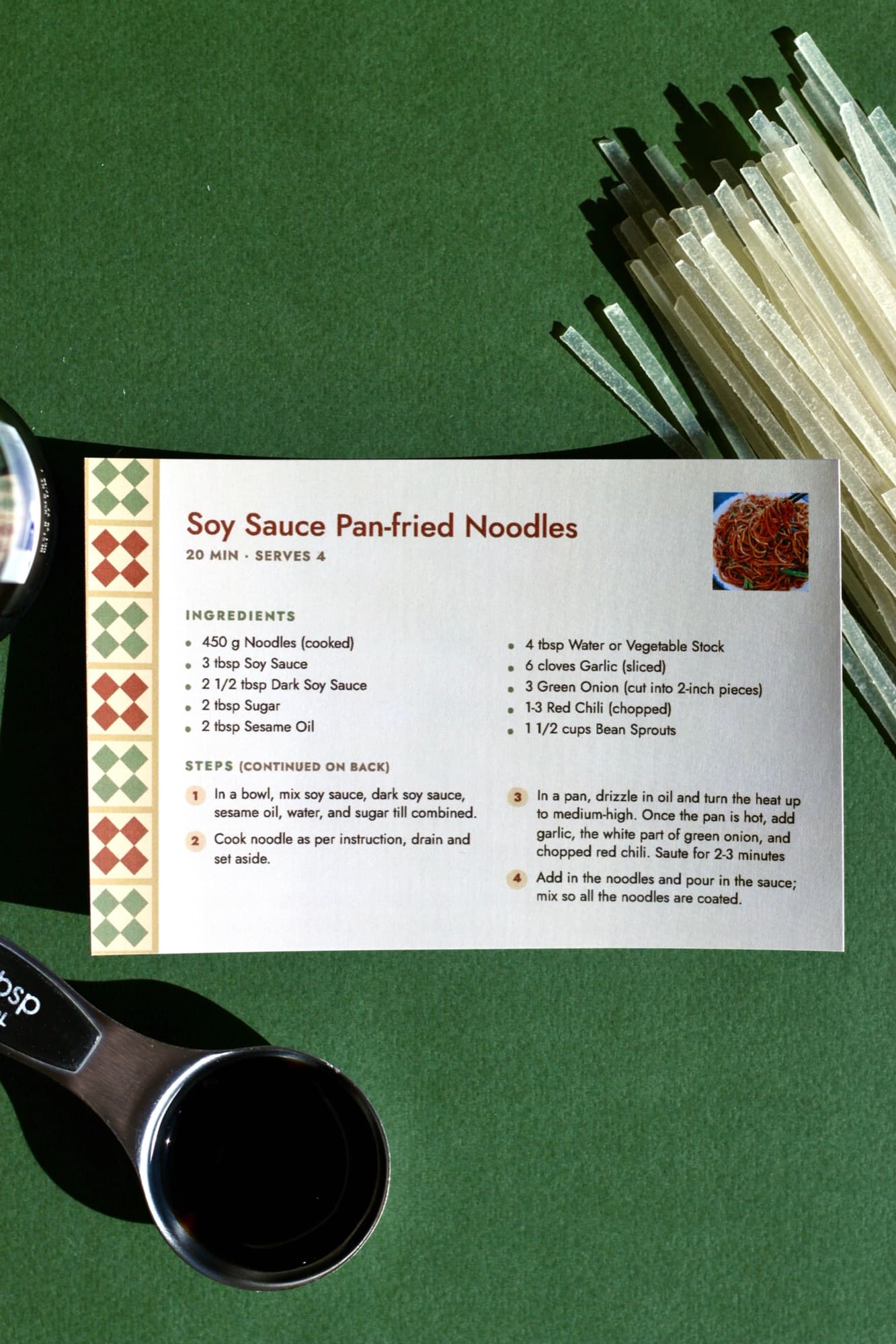A printed Soy Sauce Pan-fried Noodles card with a tiled green and red border, on a green surface beside dry rice noodles and a tablespoon of soy sauce.