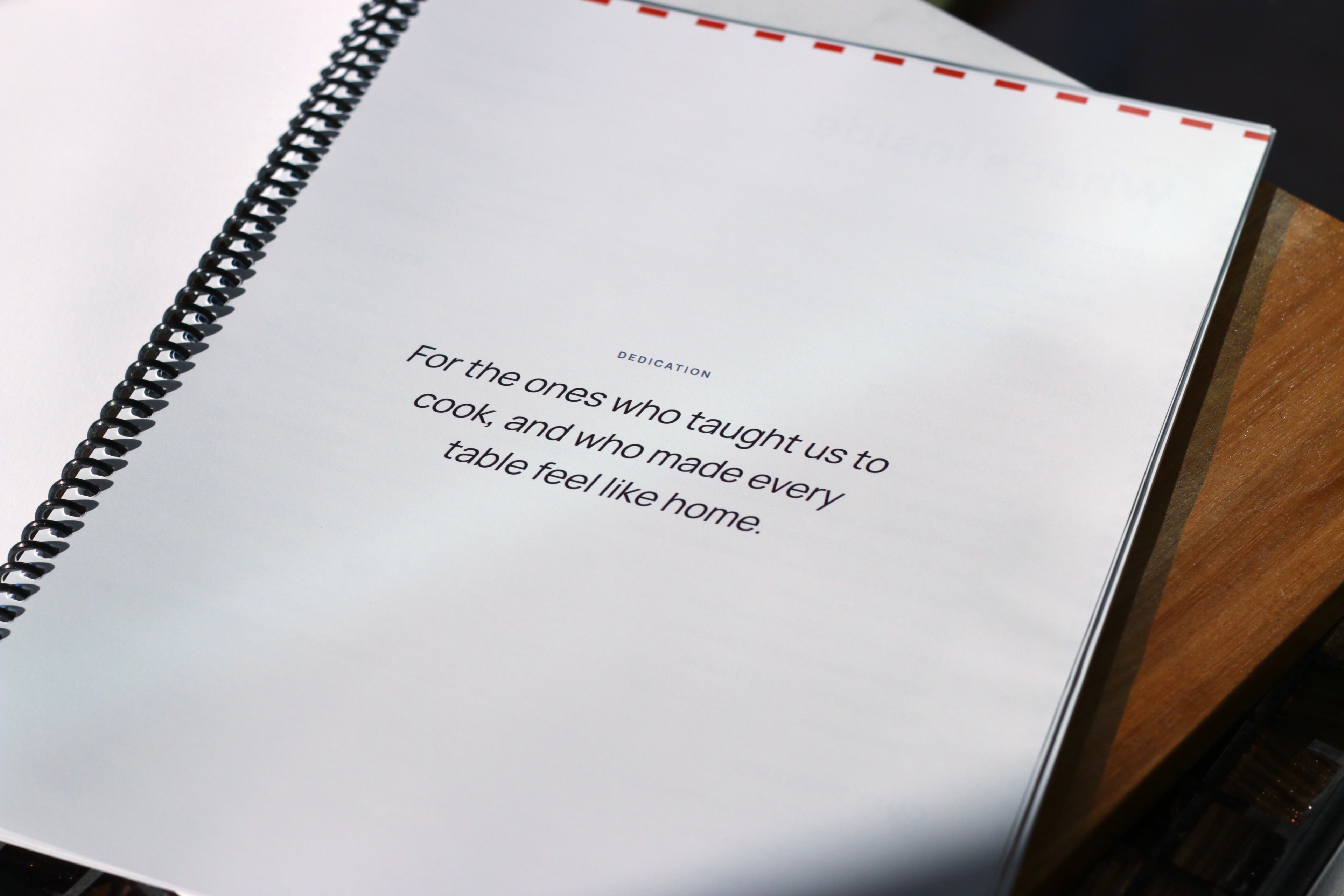 A printed, spiral-bound cookbook open to its dedication page: "For the ones who taught us to cook, and who made every table feel like home."
