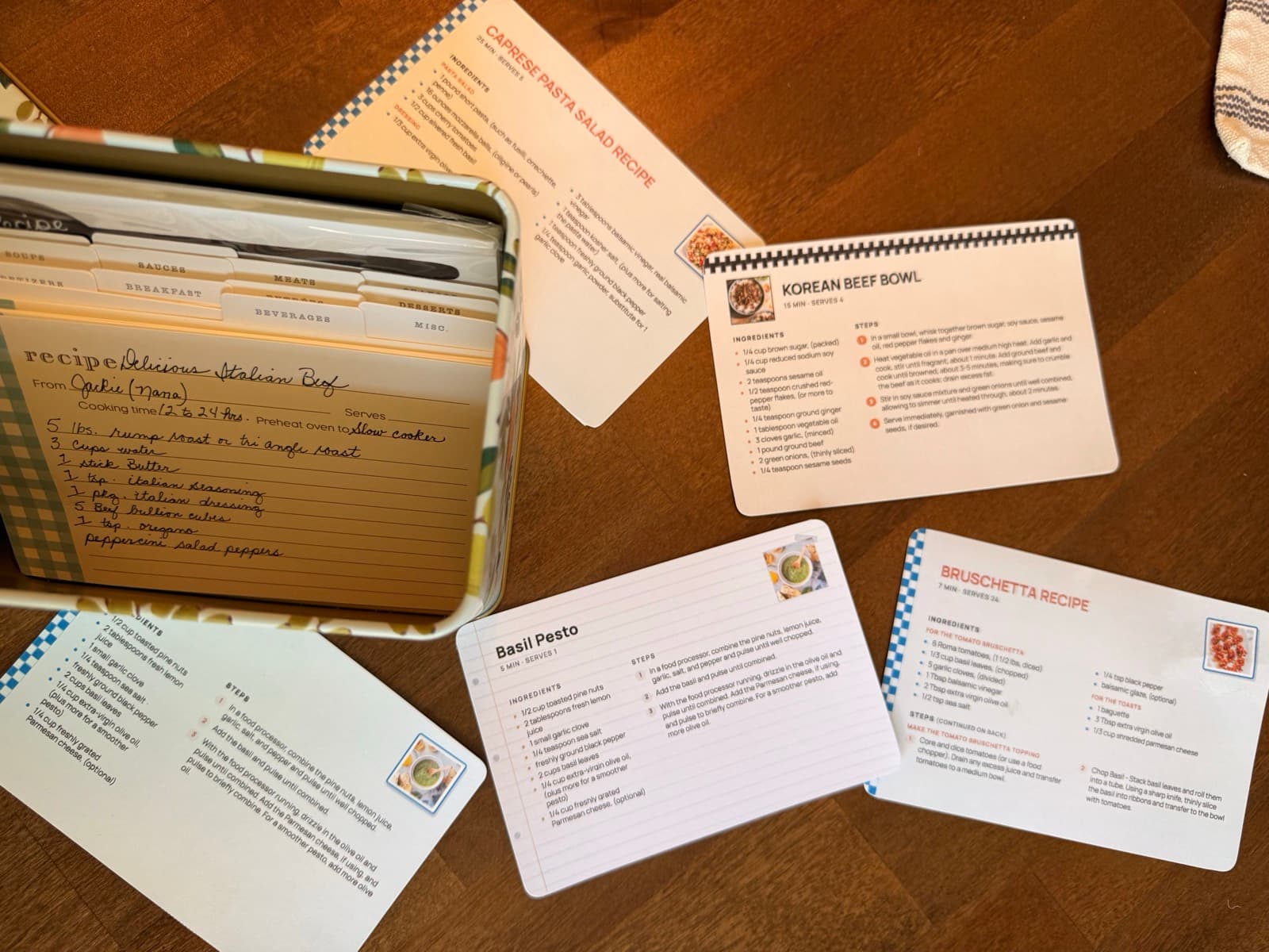Five printed recipe cards in different designs fanned across a counter beside an open recipe box.